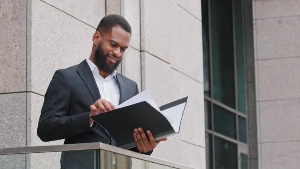 Серьезный афро-американский бизнесмен-аналитик, стоящий на балконе с документами для проверки. Молодой человек смешанной расы читает контрактные бумаги контракт, проводит исследования, готовит отчет — стоковое видео