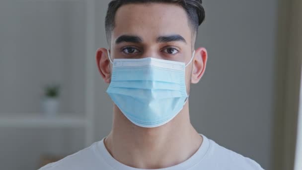 Портрет серьезного уверенного больного арабика-испанца носит защитную медицинскую маску от вируса ковид-19 на мужском лице и белую футболку, смотрящую на камеру, стоящую в помещении — стоковое видео
