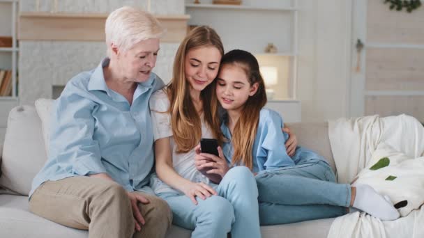 Adorable niña escolar sentado en el sofá con la madre joven y sonriendo abuela mayor madura, jugando juegos en línea utilizando aplicaciones móviles, tomando selfies divertidos juntos en casa. Familia femenina feliz — Vídeos de Stock