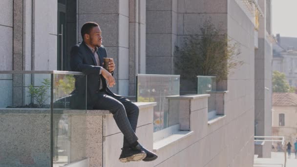 Carefree sorrindo jovem mestiço vestindo terno sentado ao ar livre perto do escritório desfrutando de pausa. confiante millennial afro-americano preto estudante trainee beber café, à espera de entrevista de emprego — Vídeo de Stock