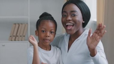 Kapalı alanda çekilmiş mutlu Afro-Amerikan genç yetişkin anne güzel koyu tenli siyah kadın ve afro küçük kız öğrenci evde oturmuş el sallıyor merhaba, selamlama hareketi, web kamerası görüntüsü