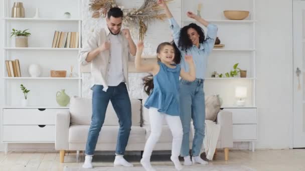 Божевільна щаслива сім'я молодих дорослих багатоетнічних батьків мама тато і милий смішний активний маленька дівчинка дочка слухає музику танцює разом, розважаючись у сучасній вітальні насолоджуючись дозвіллям — стокове відео