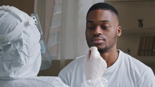 Неузнаваемый врач медсестра медицинский работник в защитной форме делает pcr тест для африканского американец больной пациент, этнический афро парень дает анализ слюны открывая рот улыбаясь, коронавируса эпидемии — стоковое видео