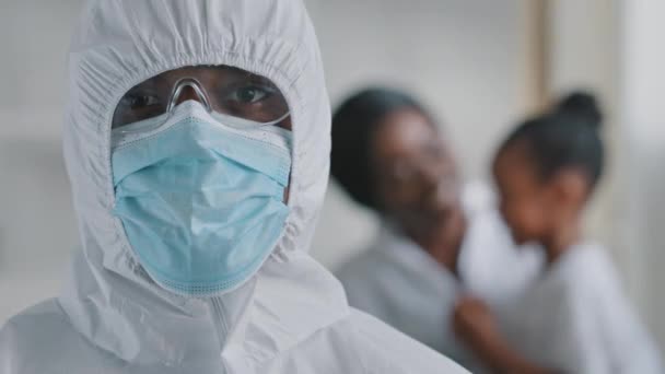 Portrait médical afro-américain médecin chirurgien regardant la caméra porte un équipement de combinaison uniforme de protection posant sur fond flou afro-américain mère de famille avec fille, femme concentrée avec enfant — Video