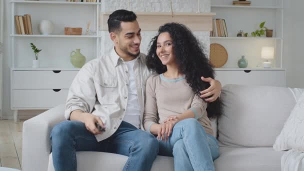 Pareja casada marido y mujer de pelo rizado hermosa mujer y hombre árabe español sentado en el sofá en casa sala de estar interior elegir canales de conmutación de programa de televisión con control remoto disfrutar de la película — Vídeos de Stock