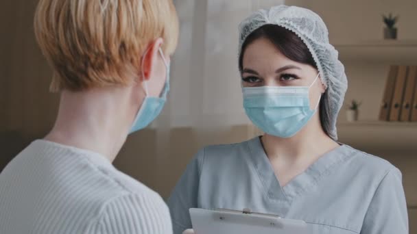 医療マスクの短い髪を持つ認識できない白人女性患者は、開業医で病院の診療所で病気の苦情の症状を書き留める女性医師看護師に話します — ストック動画