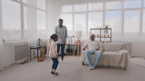 Afro amerikansk mor snurrar roterar liten dotter och leker spel med barnet medan svart far sitter på soffan i vardagsrummet. Älskade pappa hoppar upp i luften med kvinnligt barn hemma, familj tillsammans — Stockvideo