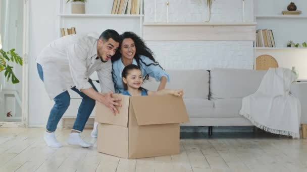 Funny active multiracial parents mom dad multiethnic family playing on moving day at new home pushing cardboard box with cute little child girl daughter kid sit inside having fun packing relocate — Stock Video