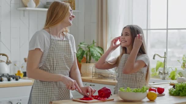 Volwassen moeder snijdt rode paprika 's in keuken koken salade met tiener meisje dochter kind neemt twee schijfjes tomaat van toepassing op oren doen alsof ze muziek te luisteren als koptelefoon geeft proberen om mam lachen — Stockvideo