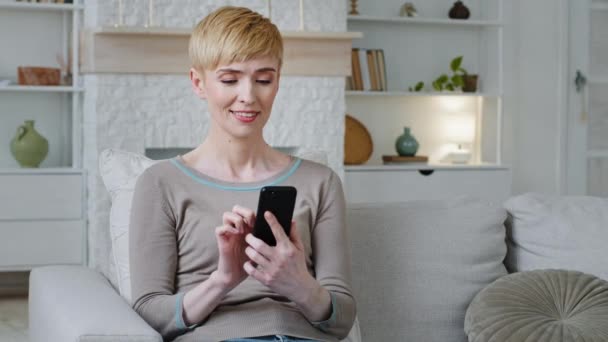 Happy relaxed woman sitting on sofa holding smartphone looking at mobilphone screen surfing social media, ellenőrzés hírek, játék mobiljátékok, sms-ek. Millenniumi hölgy töltenek időt a cellában — Stock videók