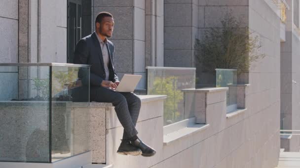 Pensativo misto empresário homem negro millennial vestindo terno sentado ao ar livre digitando no laptop, preocupado com o problema, cara pensando em tomar decisões difíceis olhando para o lado, estratégia de planejamento — Vídeo de Stock