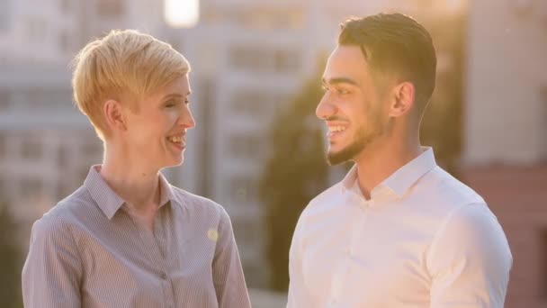 Çok ırklı iş arkadaşı çift Arap kökenli İspanyol erkek ve yetişkin beyaz kadın güneş ışığında dışarıda durmuş sohbet görüşmeleri kameraya bakıp gülümsüyor ve başlarını sallıyorlar. — Stok video