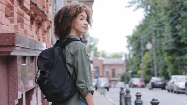 Menina americana africana atraente nova com cabelo encaracolado em pé para trás, olhando sobre o ombro fazendo gesto mão convidativo para viajar. Tipo sorrindo turista feminina com mochila convida viajar juntos — Vídeo de Stock