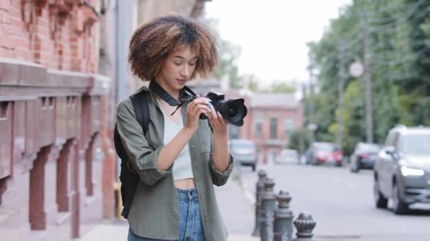 Κομψό ελκυστική σγουρά Αφροαμερικανή μαύρη γυναίκα περπάτημα δρόμο και την εξερεύνηση αξιοθέατα με την κάμερα. Νεαρή γυναίκα ταξιδιώτισσα backpacker λήψη φωτογραφιών σε φόντο θολή κτίριο τούβλο. — Αρχείο Βίντεο