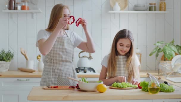 Filha adolescente ajudando a mãe com salada na cozinha em casa corta alface mãe adulta com cabelo comprido segura fatias de pimenta vermelha perto dos olhos fingindo usar óculos bebê rindo, cozinhando engraçado — Vídeo de Stock