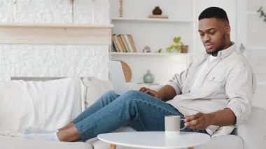Afro-Amerikan iş adamı serbest çalışan öğrenci kanepeye oturur uzaktan alışveriş için dizüstü bilgisayar kullanır internetten e-posta tarayıcıları okur internetten çay içer fincandan kahve içer mobilyaları rahat bir masaya koyar.