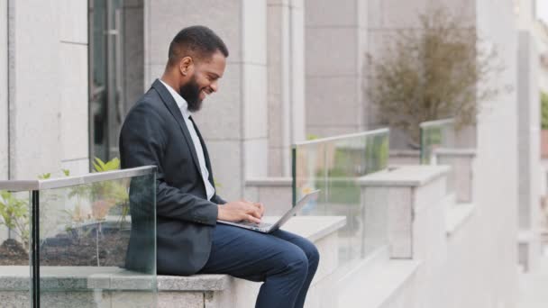 Mixte course souriant confiant jeune adulte employé corporatif masculin ou étudiant portant costume dactylographiant en plein air près du bureau. Homme d'affaires afro-américain préparant rapport d'entreprise recherche d'informations en ligne — Video