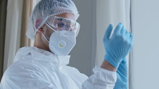 Молодой человек врач-медбрат медработник в специальной защитной форме костюма с очками и кепкой надевает резиновые голубые перчатки на руки, готовясь осмотреть пациента, стоящего у окна, глядя в камеру — стоковое видео