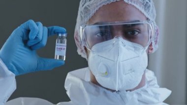 Arap adam, İspanyol doktor bilim adamı özel koruyucu üniformalı, koruyucu kıyafet gözlüklü, eldivenli Coronavirus bağışıklığı için kovid-19 aşısı olan bir ilaç şişesi taşıyan özel doktor.