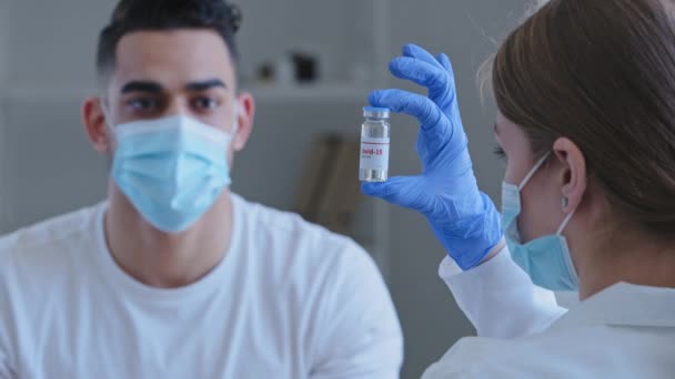 Blick von hinten kaukasische nicht wiederzuerkennende Ärztin trägt medizinische Handschuhe hält Glas mit Covid-19 Virus Impfstoff Medikament zeigt arabische Spanisch Kerl Mann Patient in Gesichtsmaske am Tisch sitzen, Impfung — Stockvideo