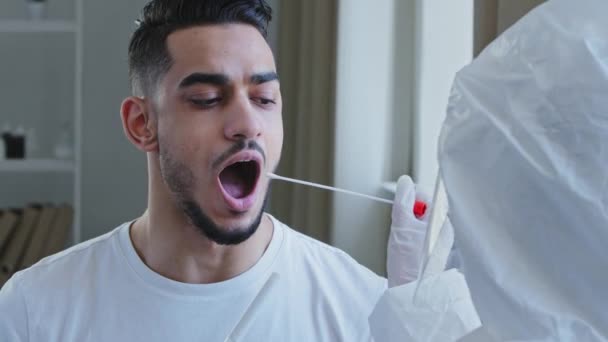 Primo piano uomo spagnolo arabo maschio paziente apre bocca dona campione di saliva per il virus dell'infezione, infermiere medico irriconoscibile facendo test PCR con attrezzature per covid-19 coronavirus al ragazzo etnico — Video Stock