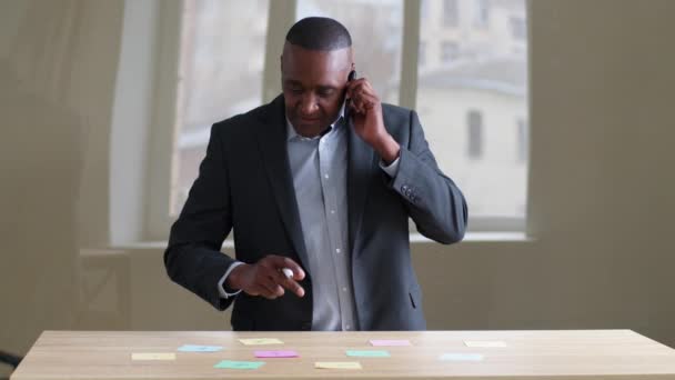 Взрослый ориентированный взрослый афроамериканец-босс мужчина, афро-лидер, носит официальный костюм, стоит за столом офиса, разговаривает по мобильному телефону с партнерами, отвечает на звонки со смартфона, пишет идеи на липких нотах — стоковое видео