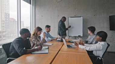 Çok ırklı iş adamları çeşitli yöneticiler çok ırklı iş adamları ofis masasında oturuyorlar beyin fırtınası yapıyorlar Afrikalı yetişkin patron beyaz tahtada yazı yazıyor.