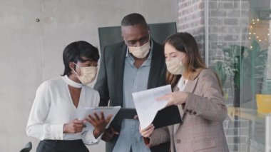 Çok ırklı iş adamları yetişkin Afrikalı erkek patron, afro siyahi kadın yönetici ve Kafkasyalı iş kadını koruyucu sağlık maskeleri takmış ellerinde belgelerle verileri kontrol ediyorlar.