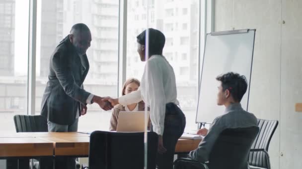 View through glass wall multiethnic business team multiracial group four people colleagues welcomes new worker boss infestor african man shaking hands making handshake partnership agreement gesture — Stock Video