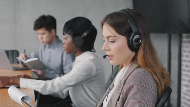 Close-up Kaukasische jonge zakenvrouw call center help lijn operator verkoper draagt headset spreekt adviseert klant op afstand het lezen van document papier zittend in de buurt van afro en aziatische collega 's — Stockvideo