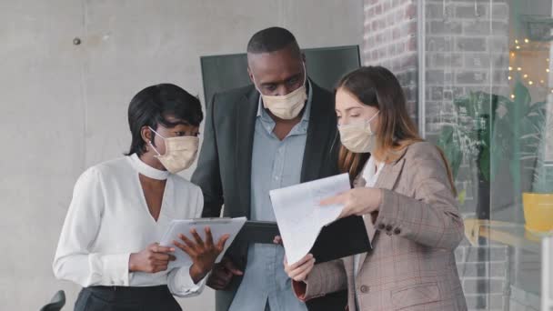 Multiracial business team adult mature african man boss, afro black woman manager and caucasian businesswoman standing in office wearing protective medical masks holding documents papers check data — Stockvideo