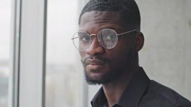Ciddi bir Afro-Amerikan iş adamı, gözlüklü, etnik siyahi, afro profesyonel bir uzman ofisinde duruyor. Gözlük takıyor ve üzgün erkek yüz ifadesi olan kameraya bakıyor.