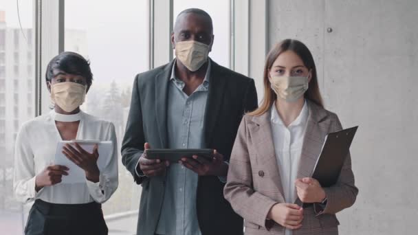 Equipo de negocios multiétnicos compañeros de grupo multirraciales compañeros de trabajo chica caucásica, mujer africana y madura jefe afro hombre con máscaras médicas protectoras posando en la oficina con documentos — Vídeos de Stock