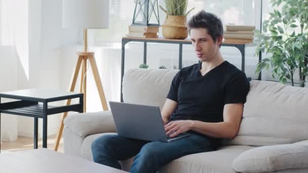 Hombre de negocios caucásico hombre milenario freelancer usa camiseta negra se sienta en el sofá utilizando el ordenador portátil para trabajar en línea de forma remota lejos de casa escribiendo chat red de navegación centrado mirando a la pantalla — Vídeo de stock