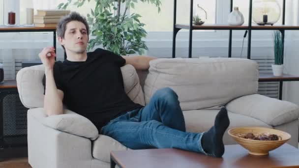 身穿黑色T恤和牛仔裤休闲服的白种人男子坐在客厅舒适的沙发上沉思着看电视节目，脚踏在木制桌子上享受周末 — 图库视频影像