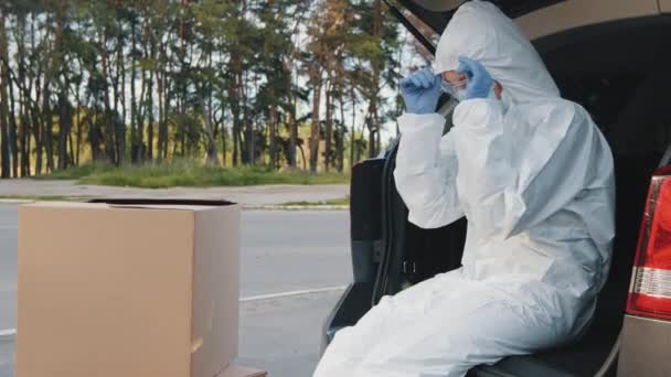 Kvinnlig kurir medicinsk arbetare bär skyddskläder latex handskar mask och transparenta glasögon korrekt huva känner värme trötthet obehag sitter i bakluckan på bilen nära kartonger tittar på kameran — Stockvideo