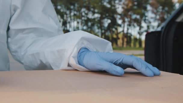 Mãos masculinas femininas de close-up estão na caixa de papelão. Correio médico irreconhecível usa roupas de proteção luvas de látex azul contém parcela com medicamentos vacinais em pé ao ar livre perto do tronco do carro — Vídeo de Stock