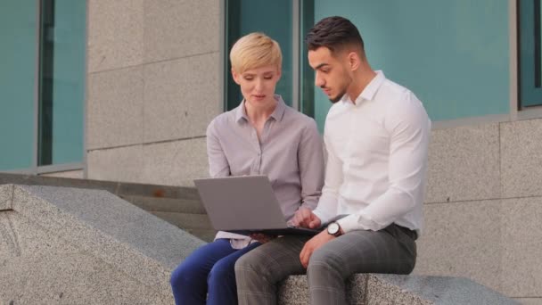 Többnemzetiségű kollégák kaukázusi nő és arab spanyol férfi ül a szabadban keres laptop sikeres győzelem. Üzletasszony számítógéppel ajánlatot tesz az üzletember győztes, hogy győzelem gesztus — Stock videók