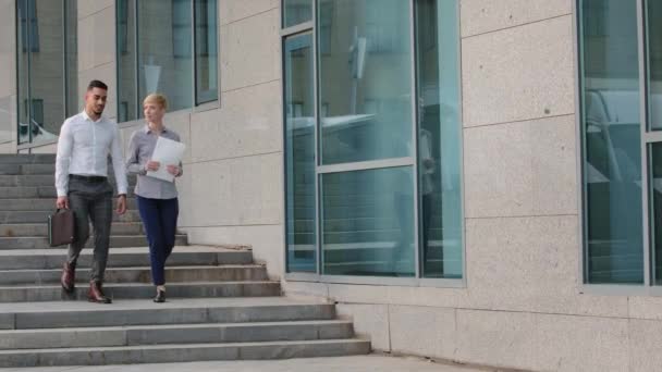 Dos compañeros de negocios multiétnicos colegas multirraciales árabe español hombre y mujer caucásica con documentos de papeles caminando por los escalones al aire libre cerca del edificio de la empresa hablar habla conversación — Vídeos de Stock