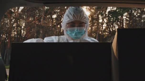 Внутрішній вигляд автомобіля жінка-кур'єр медичний працівник носить захисний одяг, маску для обличчя та прозорі окуляри стоїть біля відкритого багажника, дивлячись перевірки на обладнання для вакцин перед транспортуванням — стокове відео