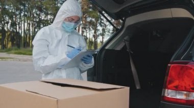 Koruyucu giysili kadın doktor kurye beyaz üniformalı lateks eldivenler araba bagajında büyük karton ilaç kutuları kağıt sayım mallarının üzerine paket sayısı yazıyor.
