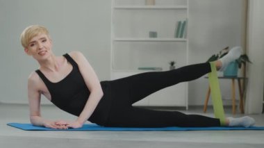 Orta yaşlı, beyaz, çevrimiçi, siyah eşofman giyen, spor yaparken gülümseyen, kameraya bakarak konuşan, elastik bant kullanarak bacaklarına ve kalçalarına egzersiz yaptıran bir kadın antrenör.
