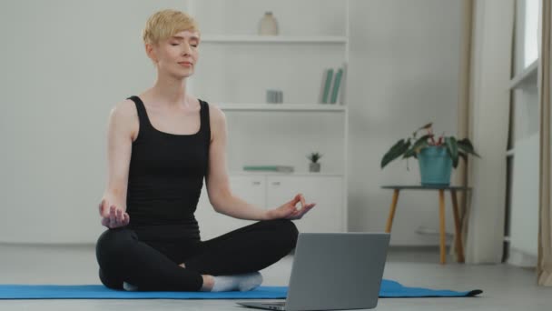 Beyaz, olgun orta yaşlı, kısa saçlı, siyah spor giysili bir kadın lotus pozisyonunda oturmuş yoga solunum egzersizleri yapıyor ve laptopuyla evde video eğitimi veriyor. — Stok video