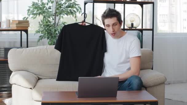 Hombre blogger de moda estilista grabación video blog en cámara web portátil. Hombre influencer de medios sociales de disparo vlog hace videollamada en casa sentado en el sofá sosteniendo negro camiseta estilista filmación tutorial — Vídeos de Stock