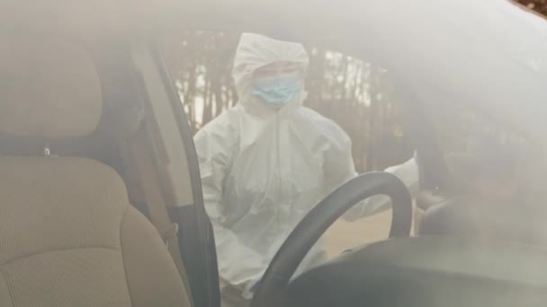 Vista attraverso il finestrino dell'auto donna irriconoscibile in tuta protettiva medica con occhiali e maschera. Medico femminile seduto nel salone automobilistico interno del veicolo che si prepara alla guida durante la pandemia coronavirica — Video Stock