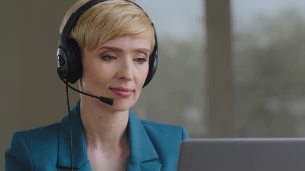 大人の成熟した40代の忙しい焦点を当てたビジネス女性オペレーターヘルプラインエージェントコールセンターサポートサービスは、カメラの笑顔を見てクライアントとオンラインチャットを作業ノートパソコン上で入力ヘッドセットを身に着けています — ストック動画