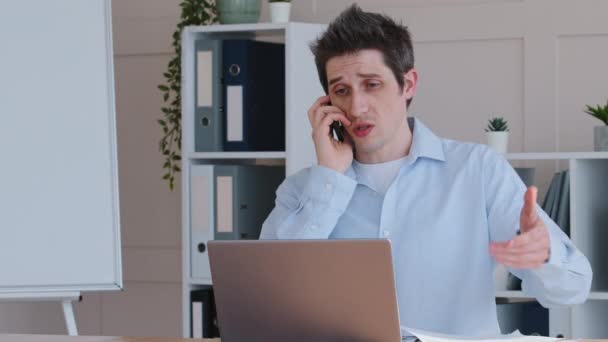 Концентрований емоційний бізнесмен чоловік кавказький менеджер чоловічої праці, що сидить за офісним столом з ноутбуком, тримає документи в руках, розмовляє мобільним телефоном, відповідає на дзвінок консультування зі смартфоном — стокове відео