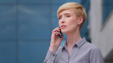 Yetişkin, 40 'lı yaşların iş kadını, olgun, akıllı, patron, sarışın, kısa saçlı, cep telefonuyla konuşan kadın portresi. Açık havada duran akıllı cep telefonuyla konuşuyor.