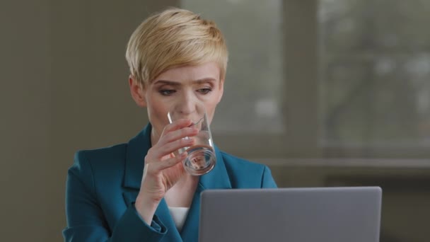 大人の成熟した40代の忙しい焦点を当てたビジネス女性短い髪は緑のジャケットを身に着けていますノートパソコンの作業を入力してオフィスのテーブルに座ってオンラインで喉の渇きを感じる透明なガラスから冷たい水を飲む — ストック動画