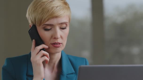 短い髪を持つ成熟した白人中高年ビジネス女性は、ノートパソコンの話して携帯電話を使用してオンライン交渉電話に答えると、会社のオフィスでテーブルに座って正式なジャケットを身に着けています — ストック動画